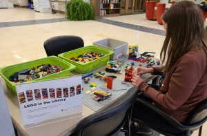 Person sitting at a table filled with lego pieces. They are building a lego creation.